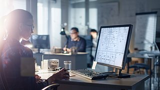 Foto, eine Frau sitzt im Büro am Schreibtisch und betrachtet technische Zeichnungen auf einem einem Computermonitor.