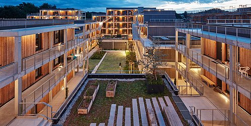 Foto, Sicht von oben auf einen Innenhof mit umliegendem Appartment-Komplex. Der Hof in der Mitte ist begrünt und mit Bodenplatten versehen. Um den Hof verläuft das mehrstöckige Gebäude.