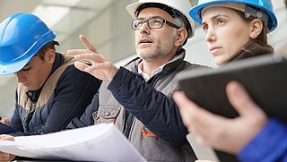 Foto, Nahaufnahme einer Gruppe von Menschen auf einer Baustelle, die einen Plan studieren. Eine Person deutet mit dem Finger aud etwas ausßerhalb des Bildauschnittes.