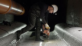 Foto, ein Arbeiter mit Helm steht in einem Tunnel und bohrt in den Boden.