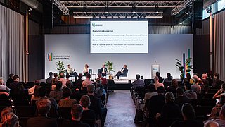 Foto, Blick aus dem Publikum auf eine Bühne auf der eine Podiumsdiskussion mit mehreren Teilnehmenden stattfindet.