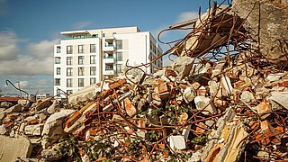 Foto, neu gebautes Gebäude im Hintergrund vor blauem Himmel, Bauschutt eines abgerissenen Gebäudes im Vordergrund.