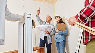 Foto, vier Personen in einem Raum auf einer Baustelle. Im Vordergrund hält eine Person ein Fensterbauteil, eine weitere Person eine Wasserwaage. Im Hintergrund studieren ein Mann und eine Frau einen Baupläne, der Mann zeigt auf etwas außerhalb des Bildauschnittes.