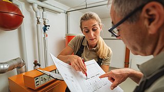 Foto, eine Monteurin und ein Monteur stehen in einem Heizungskeller und schauen gemeinsam auf ein technisches Dokument.