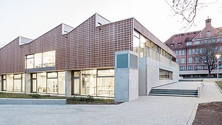 Foto, Aufnahme des neuen Schulgebäudes aus Holz und Beton. Im Hintergrund befindet sich der alte Klinkerbau des Schulgeländes. 