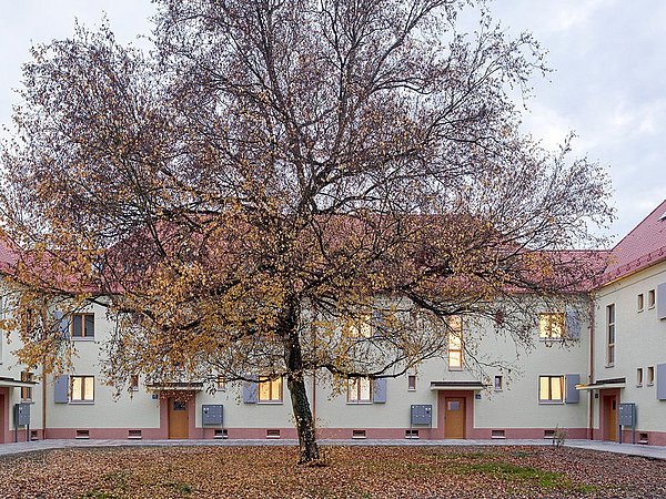 Foto, Innenhof, der von U-förmigem Gebäude umschlossen ist. In der Mitte des Hofes befindet sich ein Baum mit Herbstlaub.