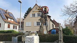 Foto, im Vordergrund befindet sich ein Lastwagen mit Baumaterialien, dahinter ein Spezialgerät zur Erdwärmebohung, im Hintergrund ein Einfamilienhaus.