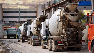 Foto, mehrere LKW-Betonmischer, die in Reihe vor einer Industrieanlage stehen.