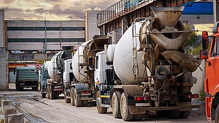 Foto, mehrere LKW-Betonmischer, die in Reihe vor einer Industrieanlage stehen.