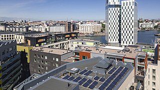 Foto, Blick von oben auf ein flaches Häuserdach mit installierten Photovoltaik-Elementen. Im Hintergrund ein modernes Hochhaus.
