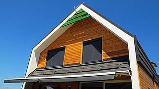 Foto, Giebelseite des Obergeschosses eines holzvertäfelten Einfamilienhauses vor blauem Himmel. 