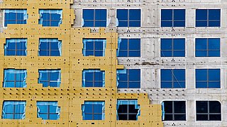 Foto, frontaler Blick auf eine mehrstöckige Neubaufassade. Die Hälfte der Fassade ist bereits mit Dämmplatten versehen, die andere Hälfte ist noch im Rohbau.