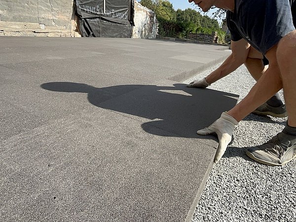 Foto, Ein Arbeiter mit Handschuhen legt ein Bodenplatte auf eine glatte Fläche und wirft einen Schatten in das helle Sonnenlicht.
