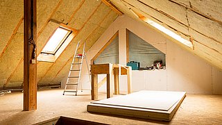 Foto, Dachboden mit schrägen Wänden, Dachfenster, Leiter, Holzstütze und gestapelten Gipskartonplatten auf dem Boden.