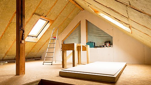 Foto, Dachboden mit schrägen Wänden, Dachfenster, Leiter, Holzstütze und gestapelten Gipskartonplatten auf dem Boden.