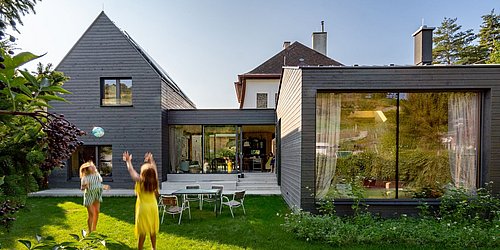 Foto, Modernes Wohnhaus mit dunkler Holzfassade, großen Glasfenstern und zwei spielenden Kindern im Garten