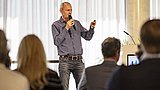 Foto, ein Redner präsentiert auf der Bühne mit einem Mikrofon, während die Zuhörer in einem gut beleuchteten Konferenzraum aufmerksam zuhören.