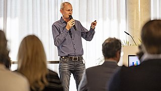 Foto, ein Redner präsentiert auf der Bühne mit einem Mikrofon, während die Zuhörer in einem gut beleuchteten Konferenzraum aufmerksam zuhören.