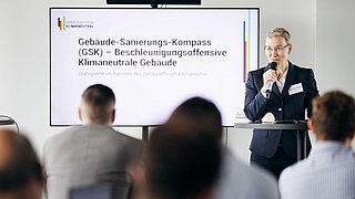 Foto, Frau im dunklen Blazer spricht mit Mikrofon vor Bildschirm mit Text zum Gebäude-Sanierungs-Kompass, Publikum im Vordergrund