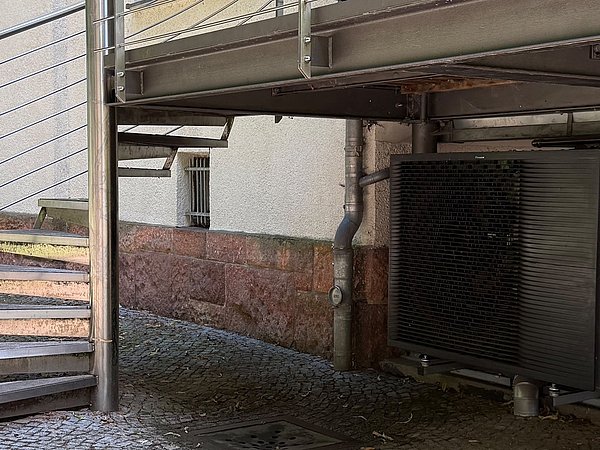 Foto, Unter einer Metalltreppe und einem erhöhten Balkon ist eine große, quaderförmige Außeneinheit einer Wärmepumpe aufgestellt. Das Gerät steht auf einem gepflasterten Boden und ist mit Rohren an die Hauswand angeschlossen. Die Fassade besteht aus hellem Putz und einem Sockel aus rötlichem Naturstein.