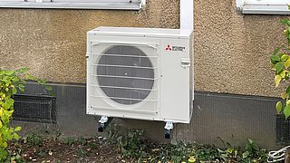 Foto, Außeneinheit einer Wärmepumpe an der Fassade eines Gebäudes mit zwei Fenstern