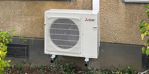 Foto, Außeneinheit einer Wärmepumpe an der Fassade eines Gebäudes mit zwei Fenstern