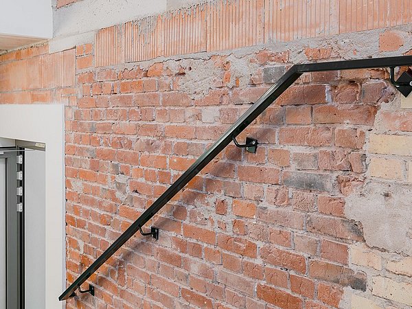 Foto, schwarzes Metallgeländer an roter Backsteinwand mit teilweise verputzten Stellen.