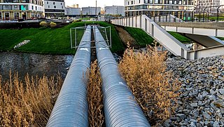 Foto, zwei große silberne Rohrleitungen verlaufen parallel über ein mit Gras bewachsenes Gelände neben einem modernen Gebäude mit mehreren Stockwerken
