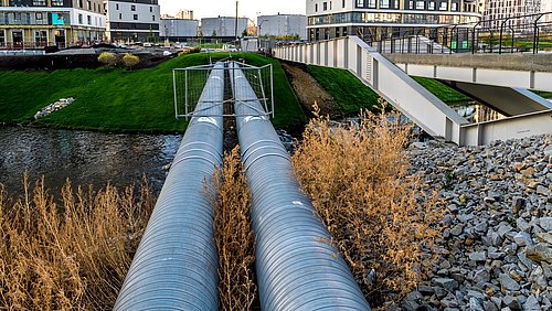 Foto, zwei große silberne Rohrleitungen verlaufen parallel über ein mit Gras bewachsenes Gelände neben einem modernen Gebäude mit mehreren Stockwerken