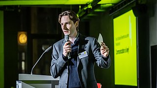 Foto, Mann in dunklem Anzug hält Mikrofon und Zettel, steht an Rednerpult vor gelber Präsentationsfolie in dunklem Raum.