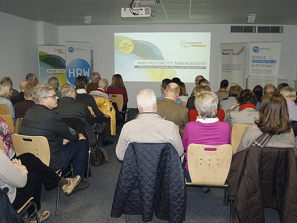 Foto, Publikum sitzt in einem Seminarraum und schaut auf eine Präsentation mit dem Titel 'Innovation City Management'