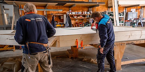 Foto, zwei Männer in Arbeitskleidung bearbeiten ein langes Holzstück in einer Werkstatt mit Regalen und Werkzeugen im Hintergrund.