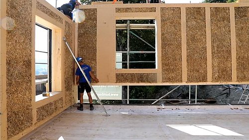 Foto, Baustelle von Gebäude mit Wänden aus Stroh. Ein Fassadenelement wird eingesetzt.