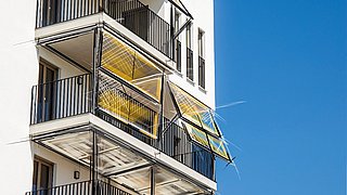 Foto, Modernes Mehrfamilienhaus mit einzigartigen gelben Markisen und schwarzen Geländern vor einem klaren blauen Himmel.