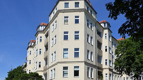 Foto, mehrstöckiges Wohngebäude mit vielen Fenstern und Erkern unter blauem Himmel und Bäumen im Vordergrund.