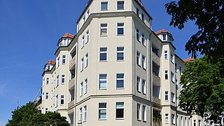 Foto, mehrstöckiges Wohngebäude mit vielen Fenstern und Erkern unter blauem Himmel und Bäumen im Vordergrund.