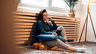 Foto, eine Frau sitzt angelehnt an einem Heizkörper und benutzt einen Tablet-Computer.