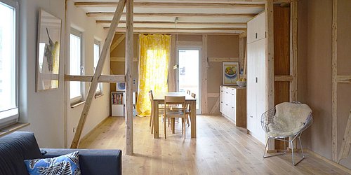 Foto, Offener Wohn- und Essbereich mit Holzbalken, Holztisch, Stühlen, gelbem Vorhang und moderner Küche im Hintergrund