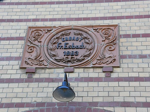 Foto, Fassadenstuck mit Verschnörkelungen. In der Mitte ist die Inschrift "Erbaut Fr. Esbach1883" zu lesen. 