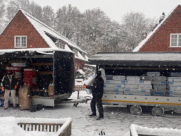 Foto, Zwei Personen laden bei Schneefall Kartons aus zwei Anhängern vor roten Backsteinhäusern mit Satteldächern.