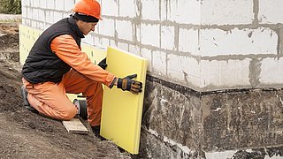 Foto, Ein Bauarbeiter kniet neben einem Hausfundament und hält eine gelbe Dämmplatte an die Außenwand.