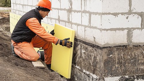Foto, Ein Bauarbeiter kniet neben einem Hausfundament und hält eine gelbe Dämmplatte an die Außenwand.