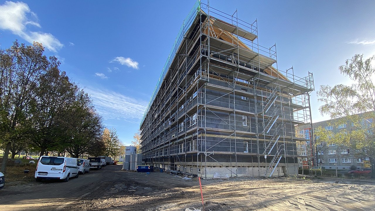 Mehrstöckiges Gebäude mit Gerüst an der Fassade, umgeben von Bäumen und geparkten Fahrzeugen unter blauem Himmel.