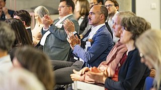 Foto, ein bunt gemischtes Publikum, das an einem modernen Veranstaltungsort sitzt, aktiv klatscht und sich während einer Präsentation oder Veranstaltung engagiert.