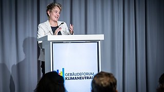 Foto, Frau in weißem Blazer spricht am Rednerpult, Publikum im Vordergrund.