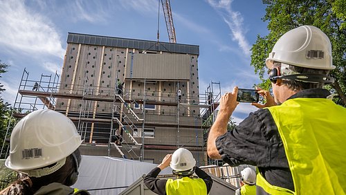 Foto, mehrere Personen mit weißen Schutzhelmen und gelben Warnwesten fotografieren ein eingerüstetes Gebäude mit Kran vor blauem Himmel.