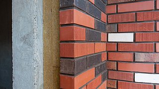 Foto, Nahaufnahme eines Wandaufbaus mit rot-braunen und weißen Ziegelsteinen, einer Dämmschicht sowie einer grauen Betonstruktur.