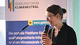 Foto, Frau mit zusammengebundenem Haar steht am Rednerpult und hält ein Mikrofon vor einem Banner mit dem Schriftzug "Gebäudeforum klimaneutral".