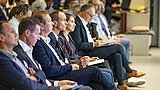 Foto, Ein konzentriertes Publikum, das in einem Konferenzraum sitzt und aufmerksam zuhört, mit Notebooks und Geräten in der Hand.