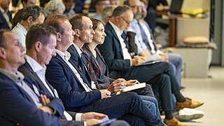 Foto, Ein konzentriertes Publikum, das in einem Konferenzraum sitzt und aufmerksam zuhört, mit Notebooks und Geräten in der Hand.